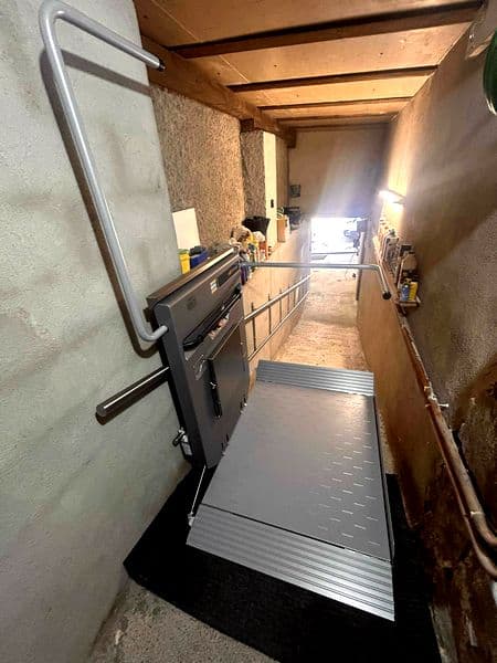 Plateforme élévatrice pour fauteuil roulant dans un couloir étroit, avec large plateau métallique antidérapant déployé et barrières de sécurité tubulaires grises.